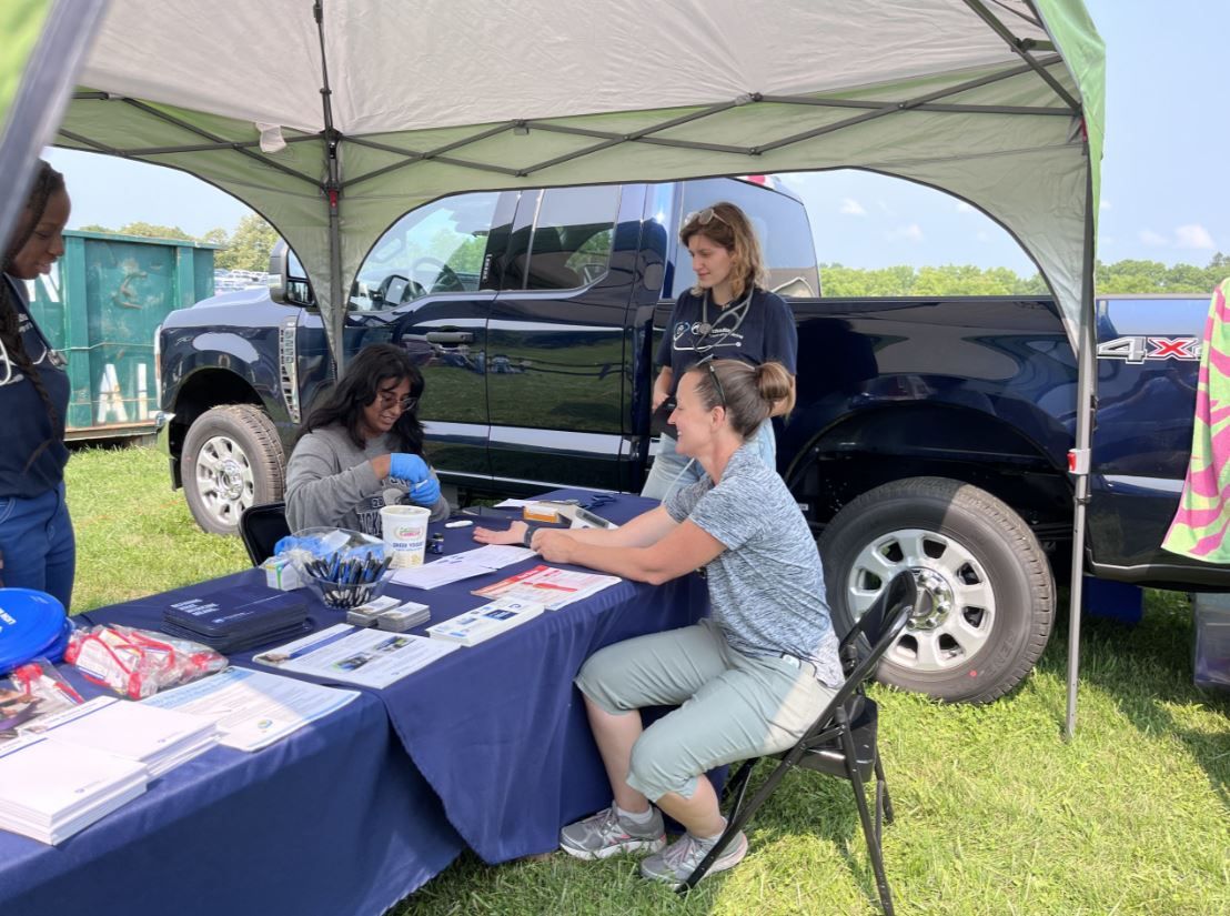 Students and faculty sit at a tent at an exhibition, providing health care screenings and education.
