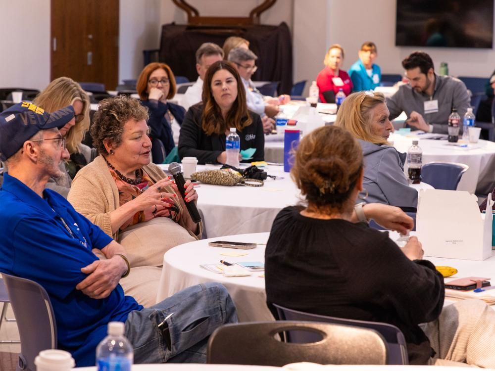 A group of people sitting at a round table discussion during Penn State CTSI’s Berks County Community-Driven Research Day. One person is speaking into a microphone, while others listen attentively.