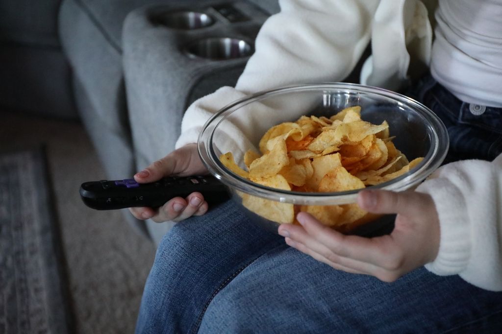 A young woman sits on a couch, holding the TV remote and a bowl of chips.